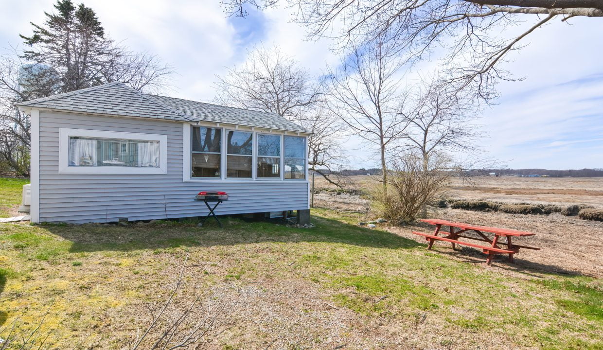 A small, gray cabin with large windows and a screened porch overlooking a barren landscape with a picnic table and a red barbecue grill outside.