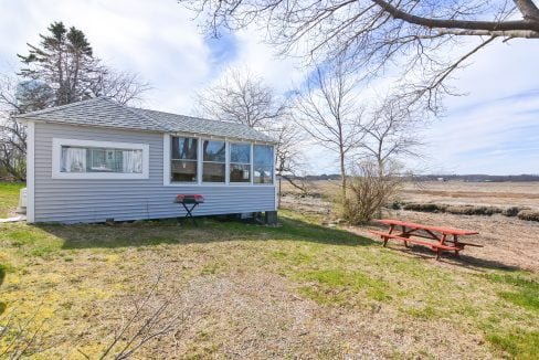 A small, gray cabin with large windows and a screened porch overlooking a barren landscape with a picnic table and a red barbecue grill outside.