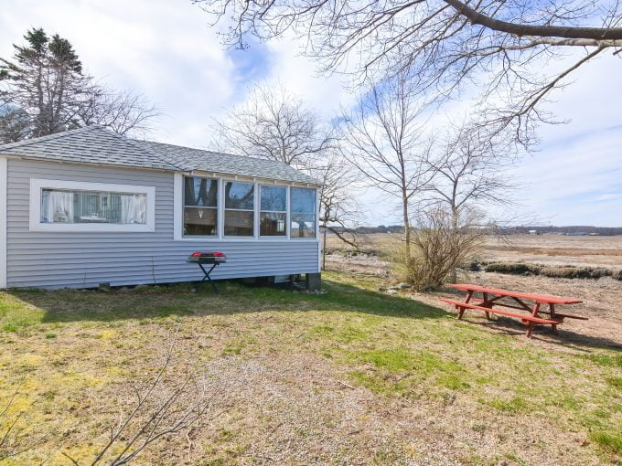 A small, gray cabin with large windows and a screened porch overlooking a barren landscape with a picnic table and a red barbecue grill outside.