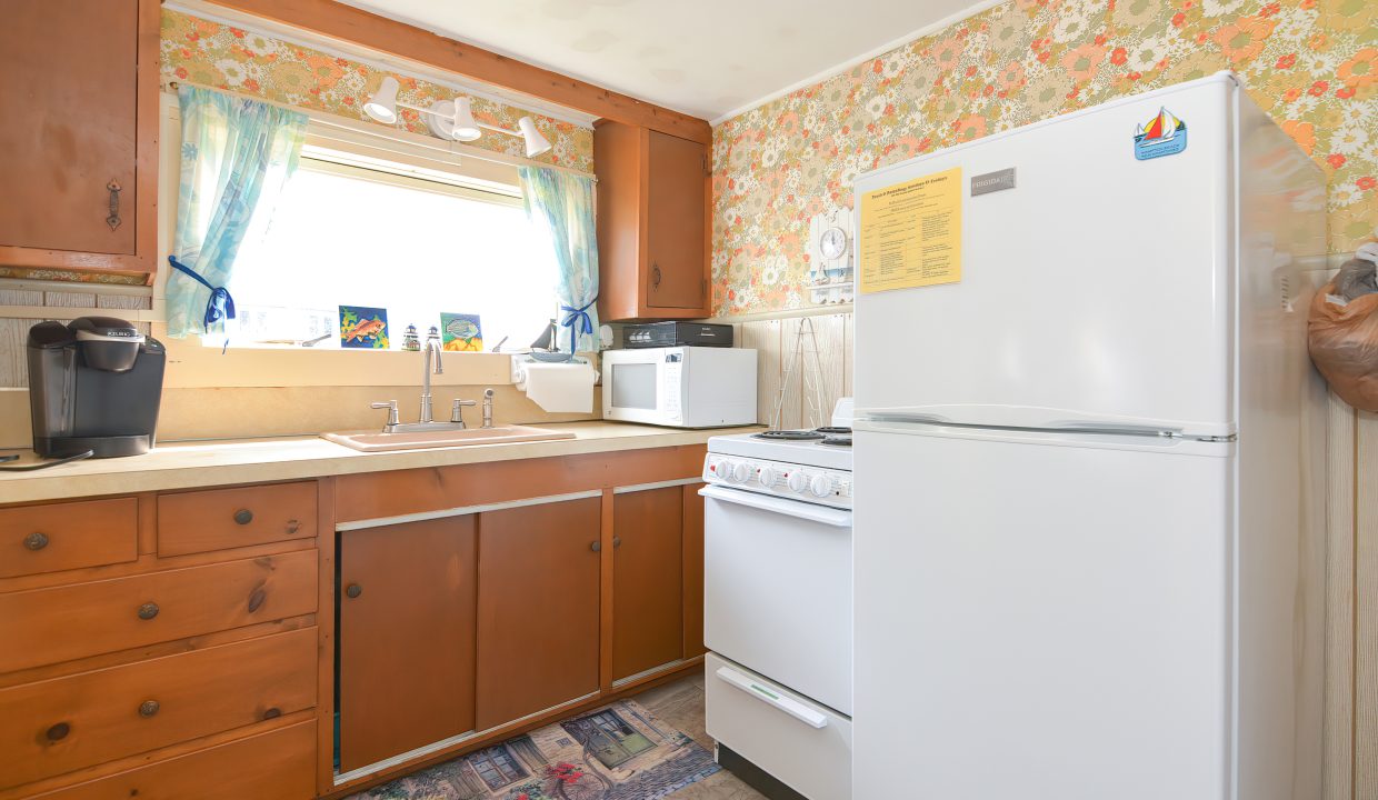 A small, retro kitchen with floral wallpaper, wooden cabinets, a window above the sink, and standard appliances including a microwave and refrigerator.