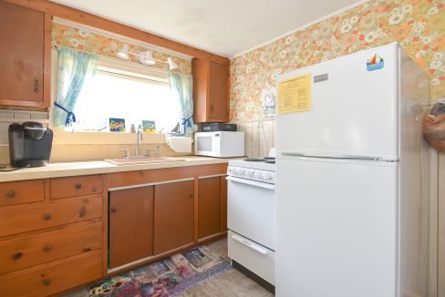 A small, retro kitchen with floral wallpaper, wooden cabinets, a window above the sink, and standard appliances including a microwave and refrigerator.