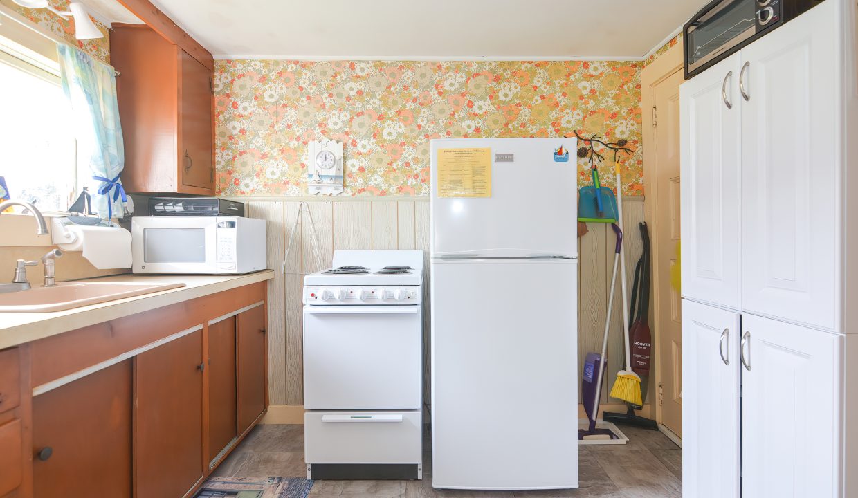 A small, vintage kitchen with floral wallpaper, wooden cabinets, and white appliances including a refrigerator, stove, and microwave.