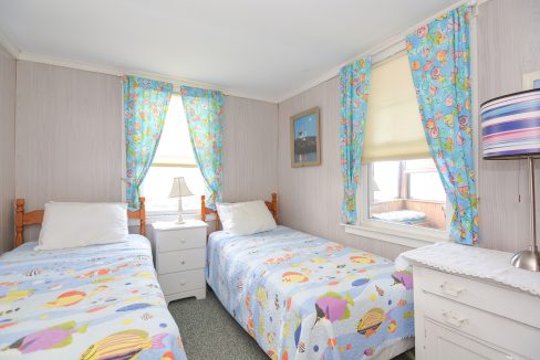 Bright, airy twin bedroom with colorful fish-themed bedding and curtains, white furniture, a small picture on the wall, and a visible window showing daylight.