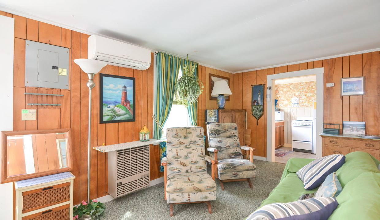 Interior of a vintage living room with wood paneling, patterned armchair, green sofa, curtain-draped windows, and doorway leading to another room.