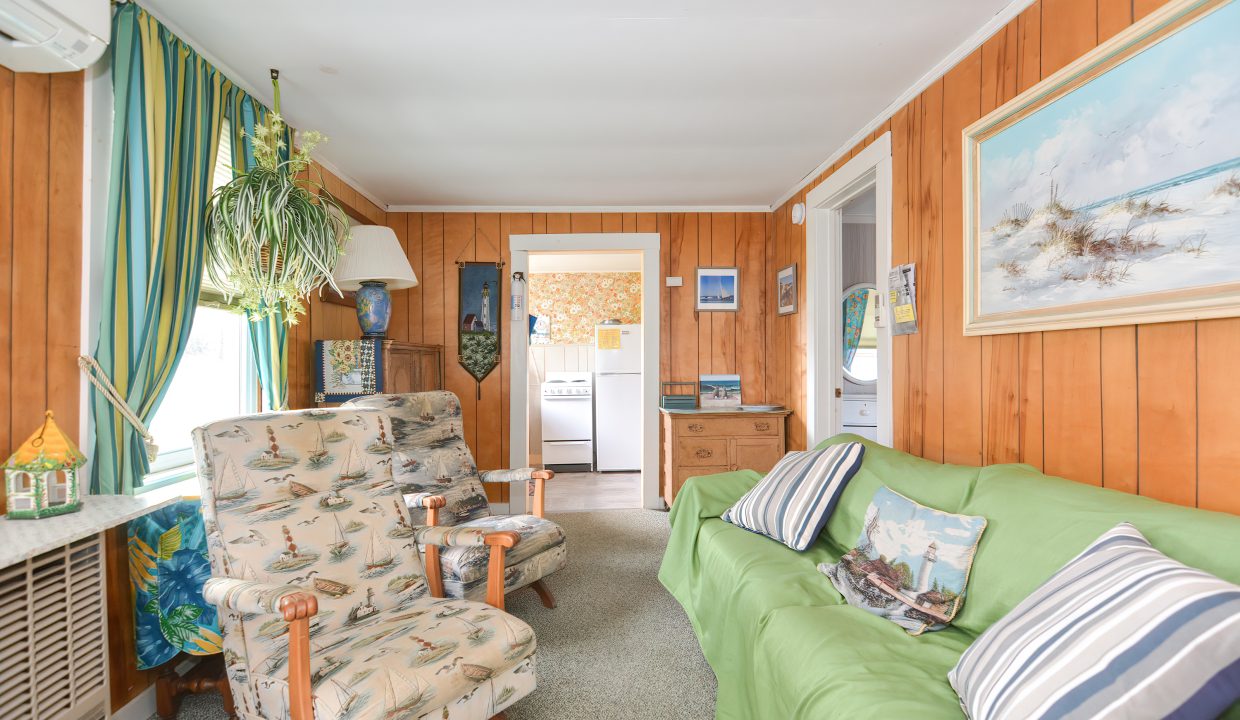 Cozy living room with green sofa, patterned armchair, wooden walls, and nautical-themed decor, leading to a small kitchen.