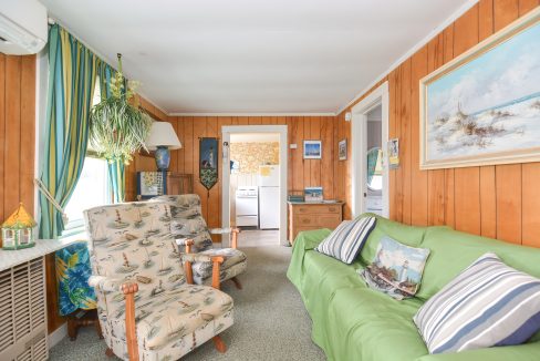 Cozy living room with green sofa, patterned armchair, wooden walls, and nautical-themed decor, leading to a small kitchen.