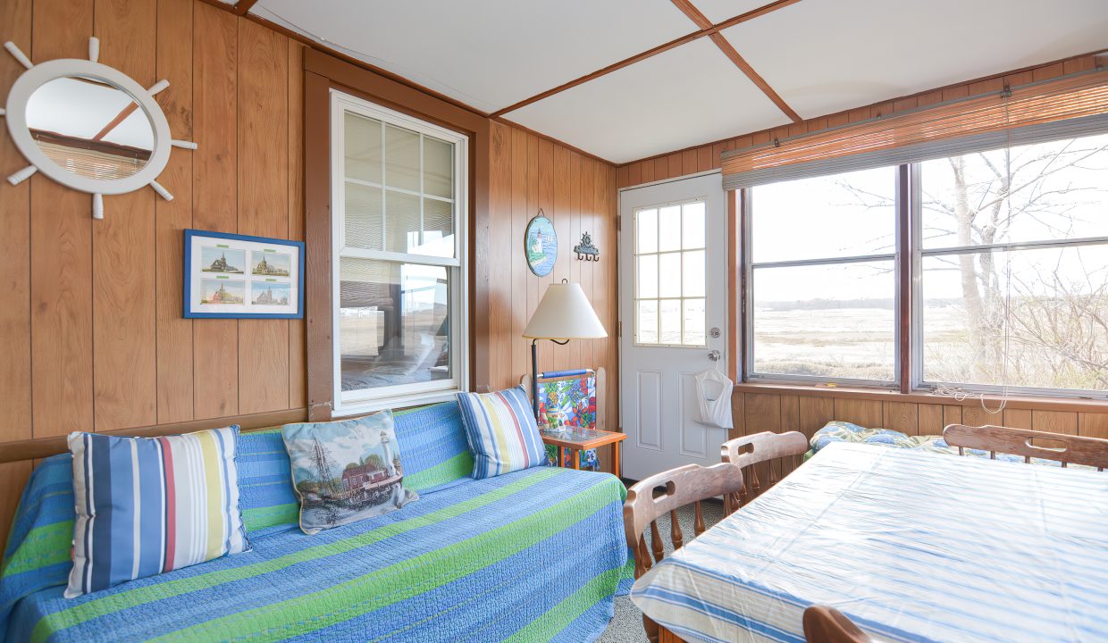 Cozy sunroom with wooden walls, large windows, patterned sofa, striped pillows, a round mirror, and a dining area with a wooden table.