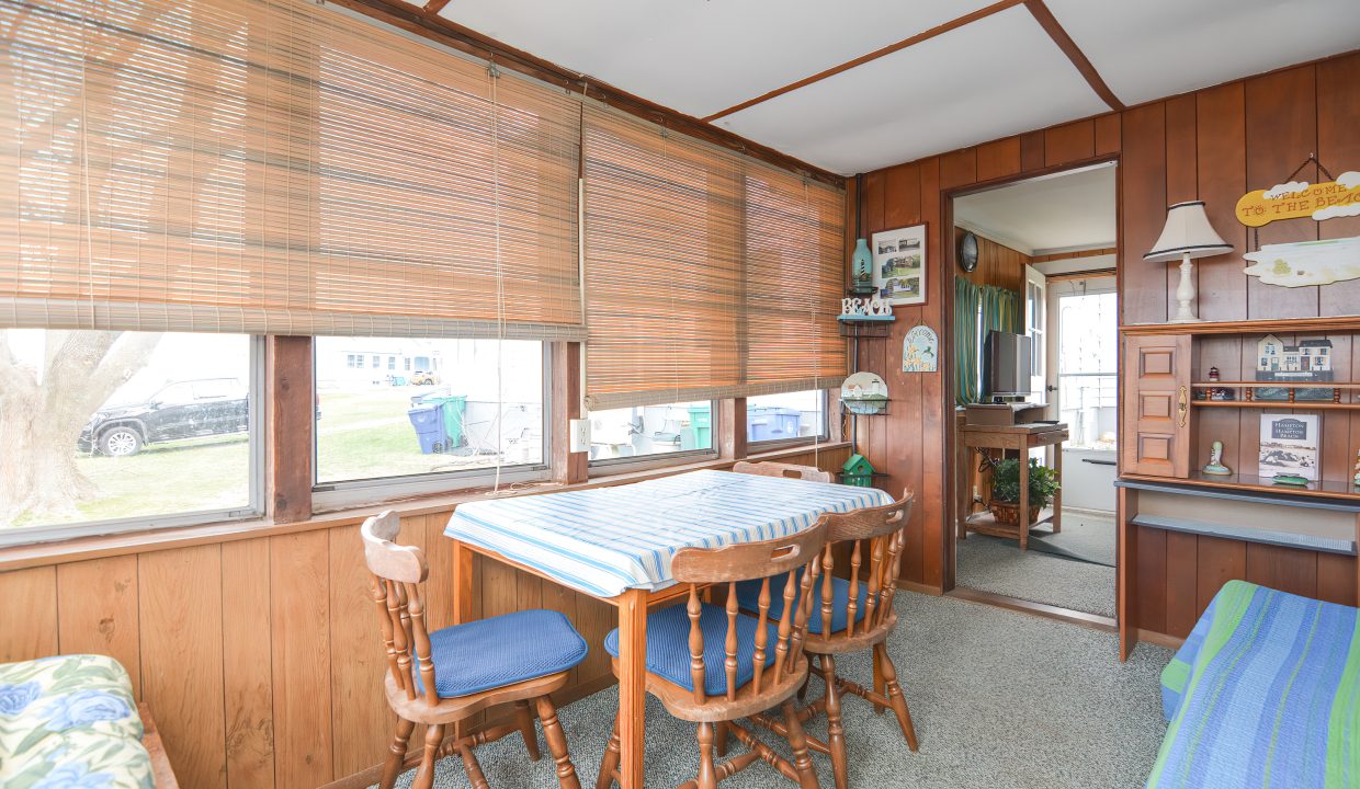 Interior of a cozy sunroom with wooden paneling, a table with chairs, blinds on large windows, and a display cabinet with various small items.