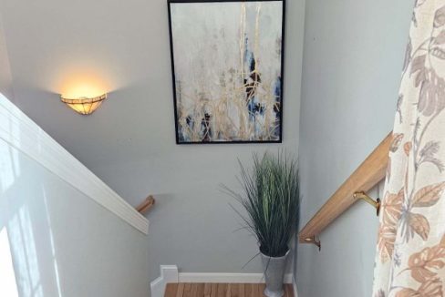 A stairway landing with an abstract painting on the wall, a wall sconce, and a decorative plant beside the wooden stairs.