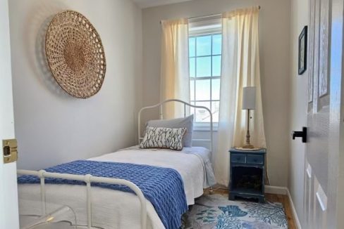 A bright, cozy bedroom with white walls featuring a single bed, a ceiling fan, patterned area rug, and a window with sheer curtains.