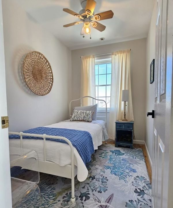 A bright, cozy bedroom with white walls featuring a single bed, a ceiling fan, patterned area rug, and a window with sheer curtains.