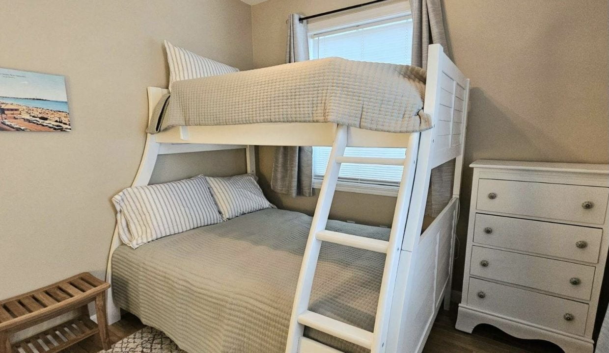 A tidy room with a white wooden bunk bed, striped bedding, a dresser, and a small window with curtains.