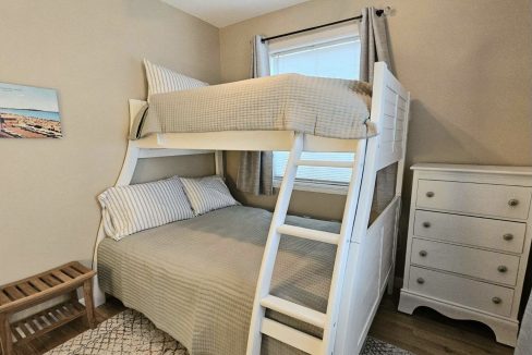 A tidy room with a white wooden bunk bed, striped bedding, a dresser, and a small window with curtains.