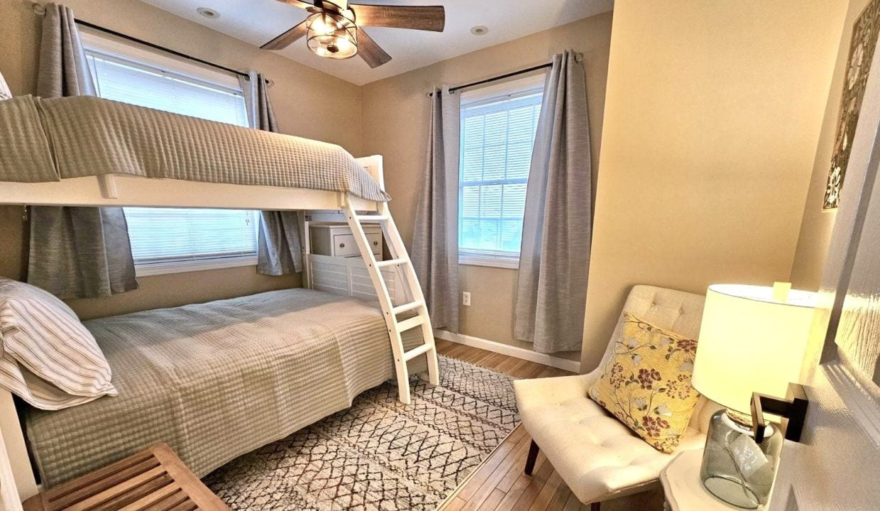 A cozy bedroom featuring a bunk bed with a ladder, matching textiles, a ceiling fan, and a lit table lamp beside a window with curtains.