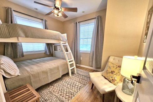 A cozy bedroom featuring a bunk bed with a ladder, matching textiles, a ceiling fan, and a lit table lamp beside a window with curtains.