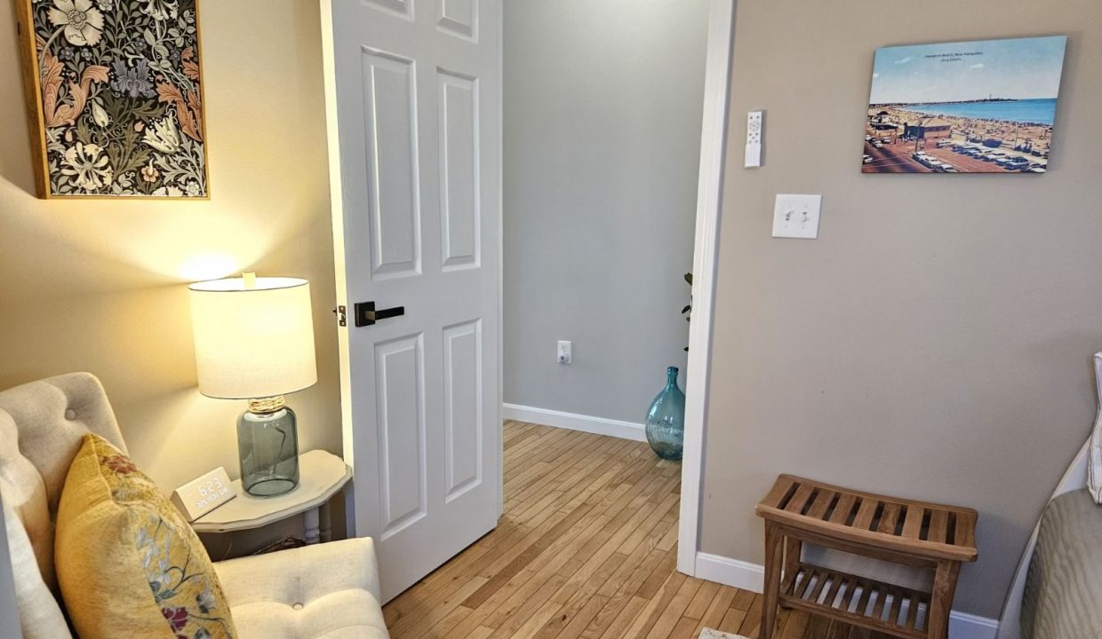 A cozy corner in a room with hardwood floors, featuring a white couch with a floral cushion, a side table with a lamp, a decorative vase on the floor, framed artwork on the walls, and a closed white door.