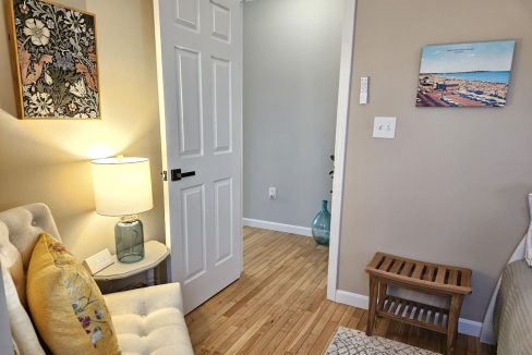 A cozy corner in a room with hardwood floors, featuring a white couch with a floral cushion, a side table with a lamp, a decorative vase on the floor, framed artwork on the walls, and a closed white door.