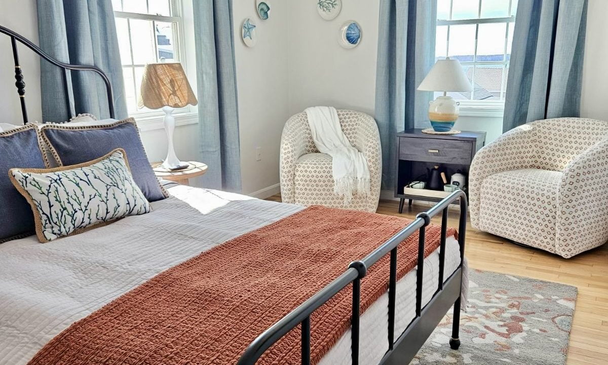 A bright, well-lit bedroom with a metal frame bed, terracotta bedspread, hardwood floor, and blue curtains.