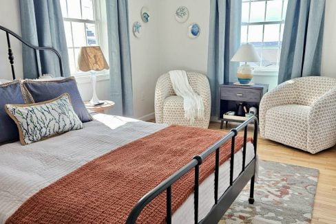 A bright, well-lit bedroom with a metal frame bed, terracotta bedspread, hardwood floor, and blue curtains.