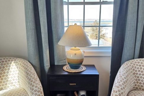 A cozy seating area by a window with a view, featuring two patterned chairs flanking a side table with a lamp.