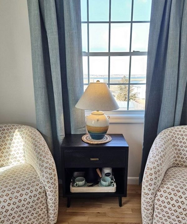 A cozy seating area by a window with a view, featuring two patterned chairs flanking a side table with a lamp.