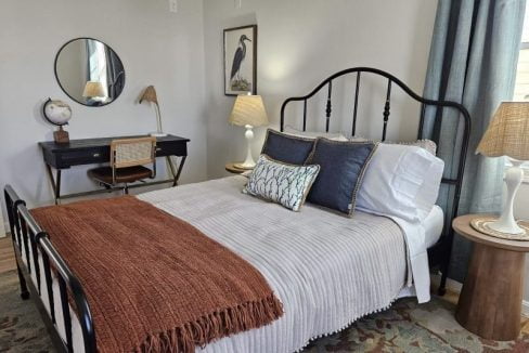 A neatly arranged bedroom with a metal bed frame, decorative pillows, a terracotta throw blanket, two bedside tables with lamps, and a writing desk with a round mirror above it.