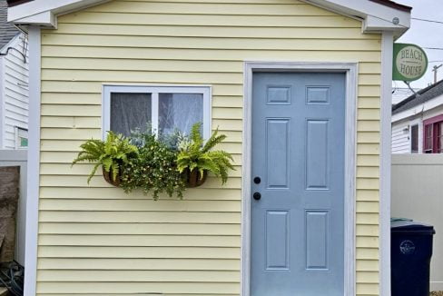 A small yellow house with a blue door and a window box containing green plants.