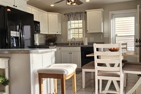 A well-lit kitchen with white cabinetry, modern appliances, and a breakfast bar with stools under a ceiling fan.