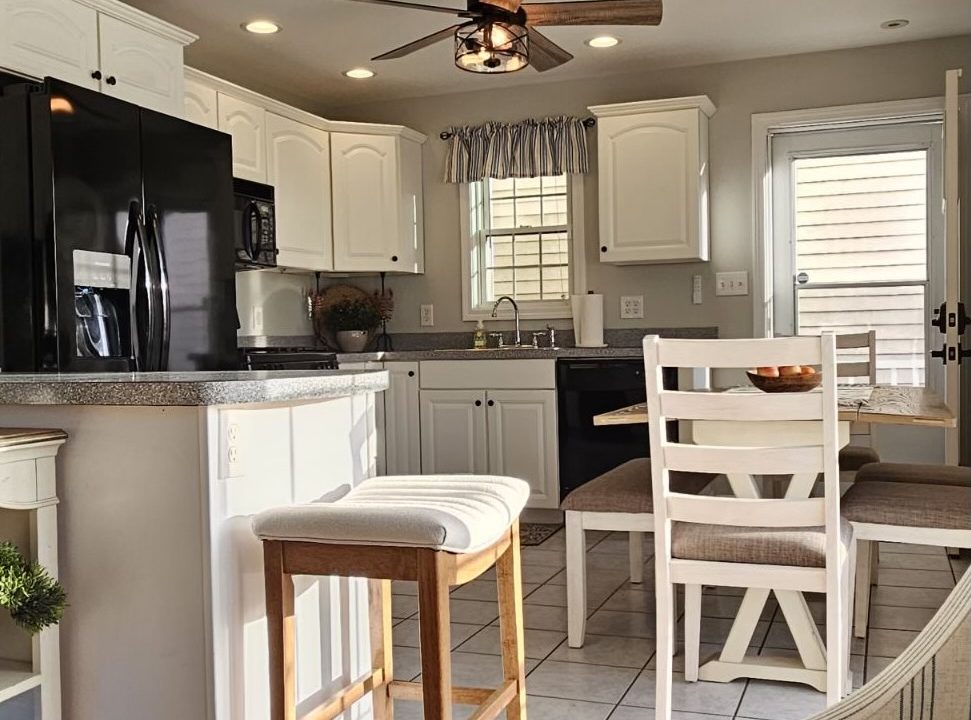 A well-lit kitchen with white cabinetry, modern appliances, and a breakfast bar with stools under a ceiling fan.