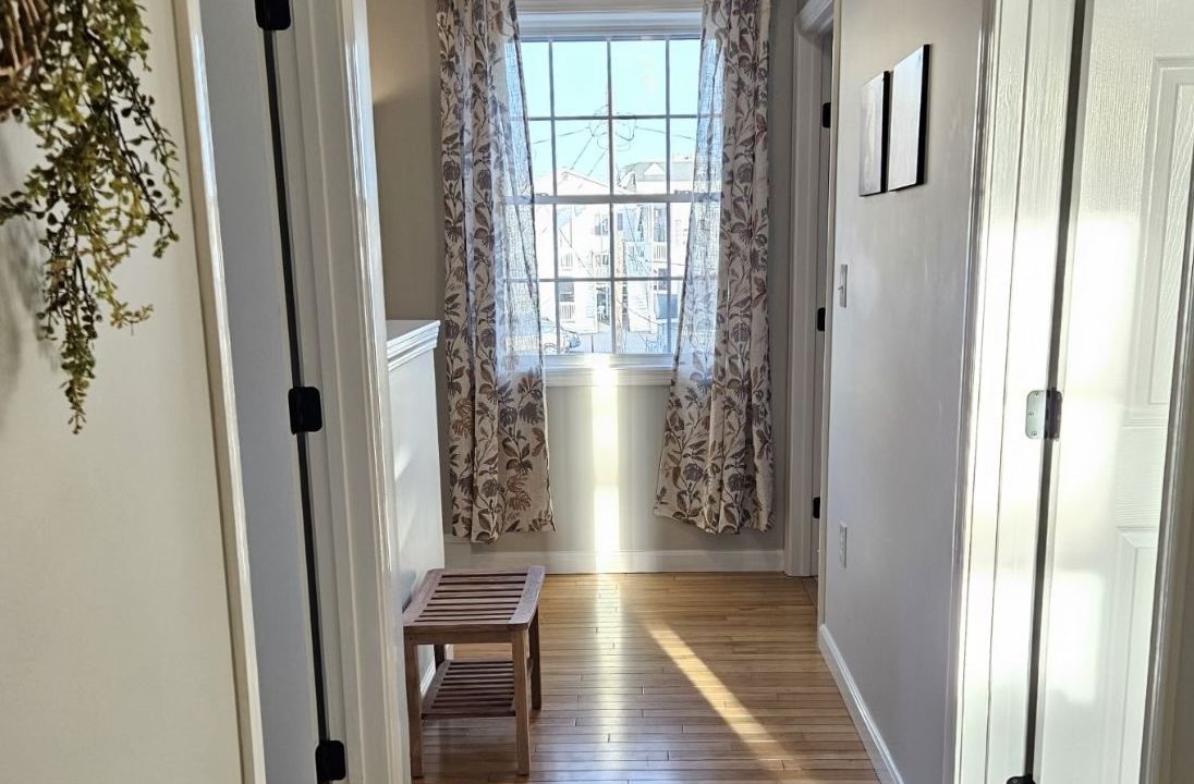 A bright hallway with wooden flooring, floral curtains on a window, and a small bench under the window.