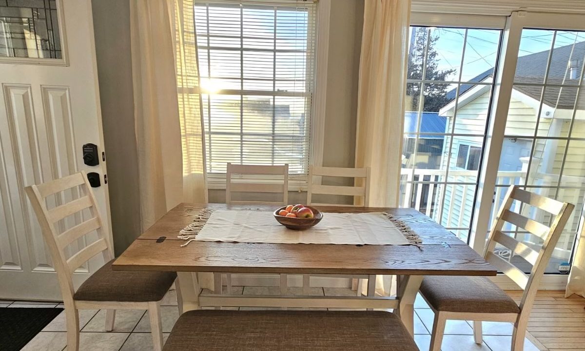 A bright dining room with a wooden table set, light curtains, and a view of a patio area through sliding doors.