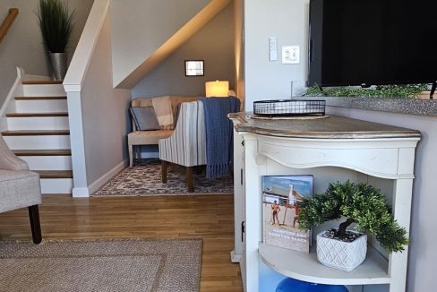 A cozy living room corner with a staircase, a lamp on a side table, and a television set, featuring decorative elements and a neutral color palette.