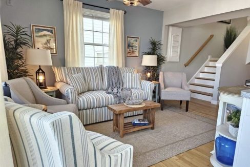 A cozy, nautical-themed living room with striped sofas, a wooden coffee table, and a staircase leading to the upper level.