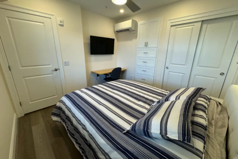 A neatly arranged bedroom with a striped bedspread, a mounted television, and a small work desk.