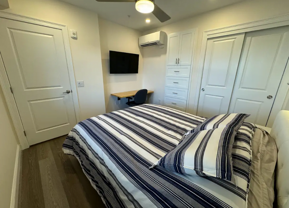 A neatly arranged bedroom with a striped bedspread, a mounted television, and a small work desk.