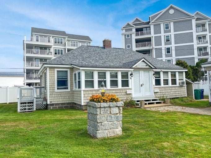 Small traditional cottage with white siding and shingles, surrounded by a grassy area and larger modern buildings in the background.