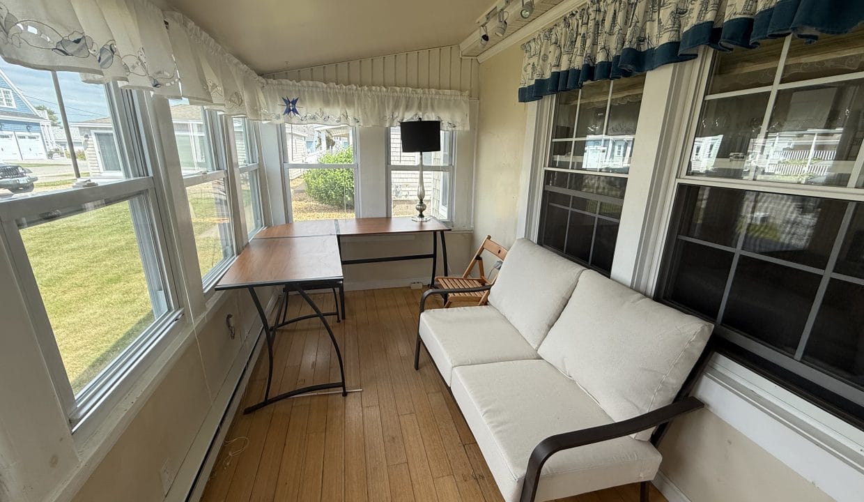 A sunroom with large windows, a white sofa, a wooden chair, a lamp, and two tables with light wood floors and nautical-themed curtains.
