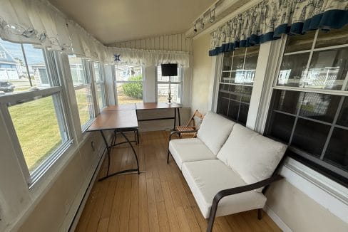 A sunroom with large windows, a white sofa, a wooden chair, a lamp, and two tables with light wood floors and nautical-themed curtains.