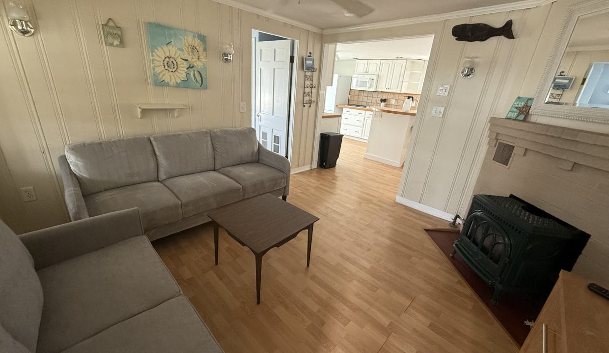 A small living room with two gray couches, a wooden coffee table, a fireplace, and a view into a bright kitchen with white cabinets.
