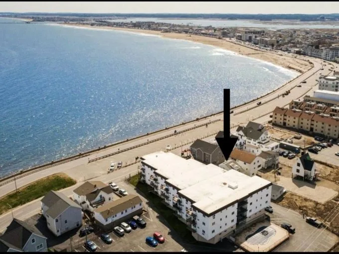 A coastal town with a beach, showing buildings near the shore. An arrow points to a specific building. Cars are parked along a road by the beachfront.
