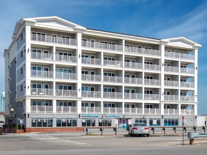 A multi-story building with balconies and a business on the ground floor, situated near a street with parked cars under a clear blue sky.