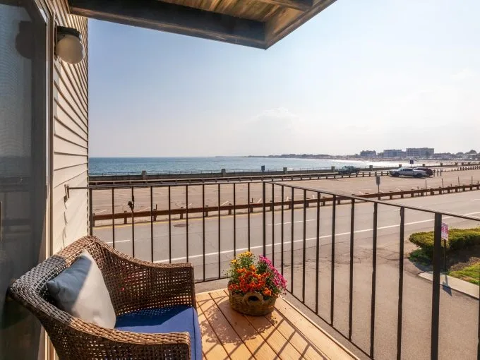 A small balcony with a wicker chair and a potted plant overlooks a road, beach, and ocean under a clear sky.