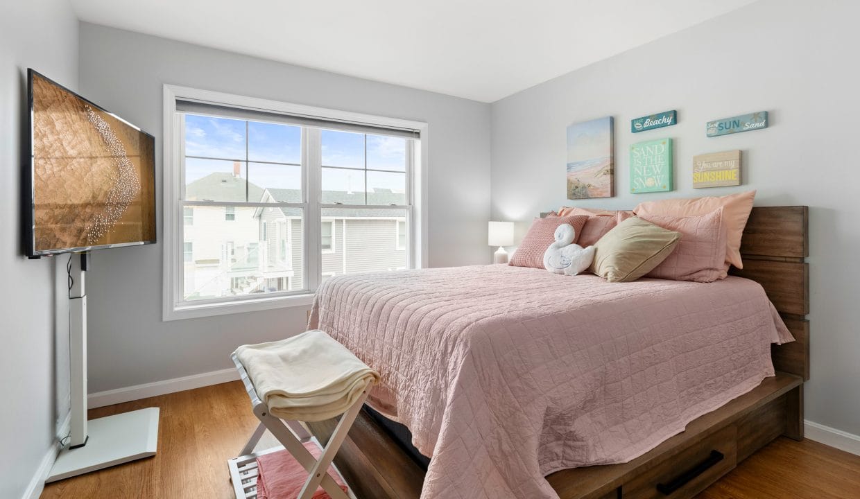 A neatly made bed with pink bedding in a bright bedroom, featuring a large window, wall art, a wooden floor, and a TV mounted on the left wall.