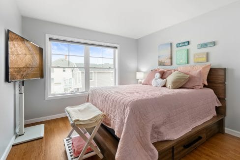 A neatly made bed with pink bedding in a bright bedroom, featuring a large window, wall art, a wooden floor, and a TV mounted on the left wall.