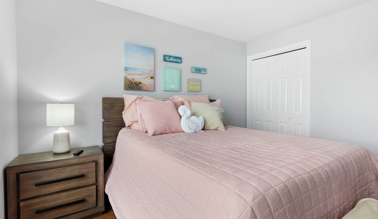 A neatly made bed with a pink quilt, decorative pillows, and a towel folded like a swan, beside a wooden nightstand with a lamp and remote; wall art hangs above.