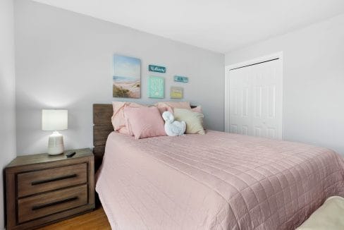 A neatly made bed with a pink quilt, decorative pillows, and a towel folded like a swan, beside a wooden nightstand with a lamp and remote; wall art hangs above.