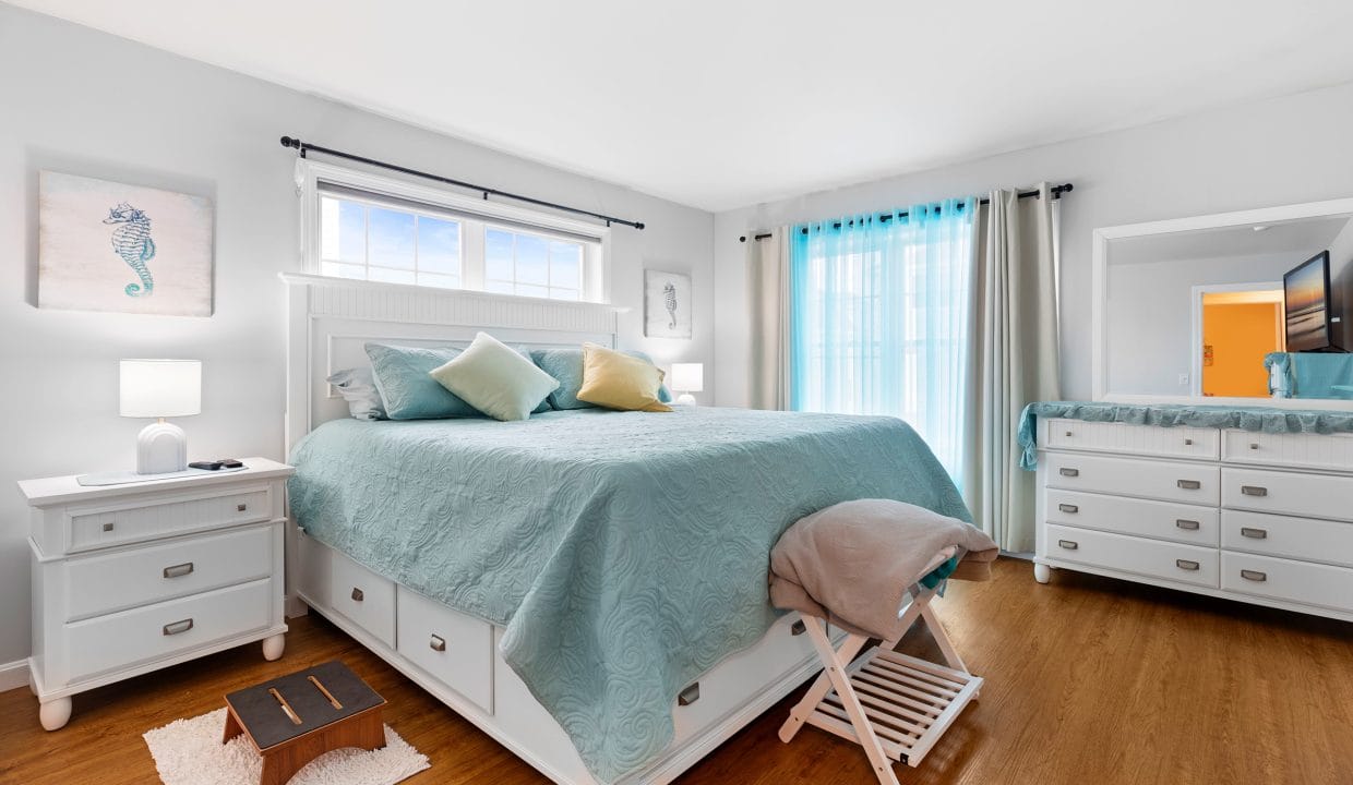Bright bedroom with a large bed, white furniture, bamboo flooring, sea-themed wall art, and sheer blue curtains over a window.