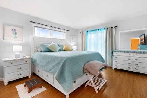 Bright bedroom with a large bed, white furniture, bamboo flooring, sea-themed wall art, and sheer blue curtains over a window.
