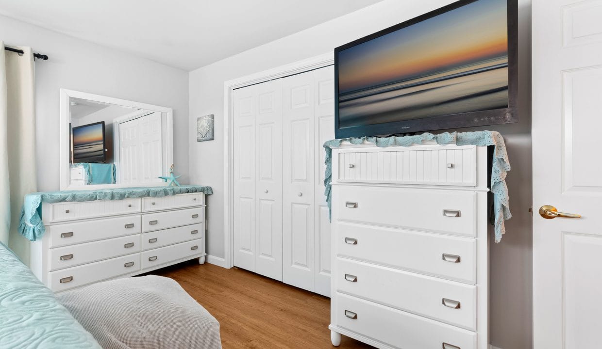 A bedroom with white furniture, including a dresser with a TV on top, a mirrored vanity, hardwood floors, and neutral walls.