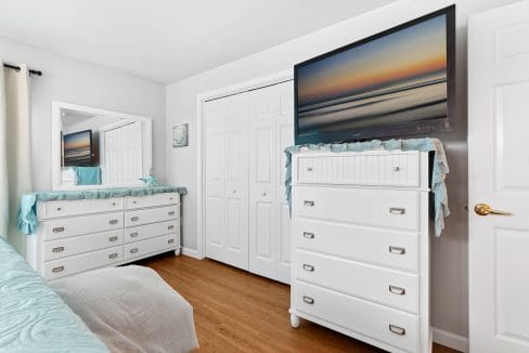 A bedroom with white furniture, including a dresser with a TV on top, a mirrored vanity, hardwood floors, and neutral walls.